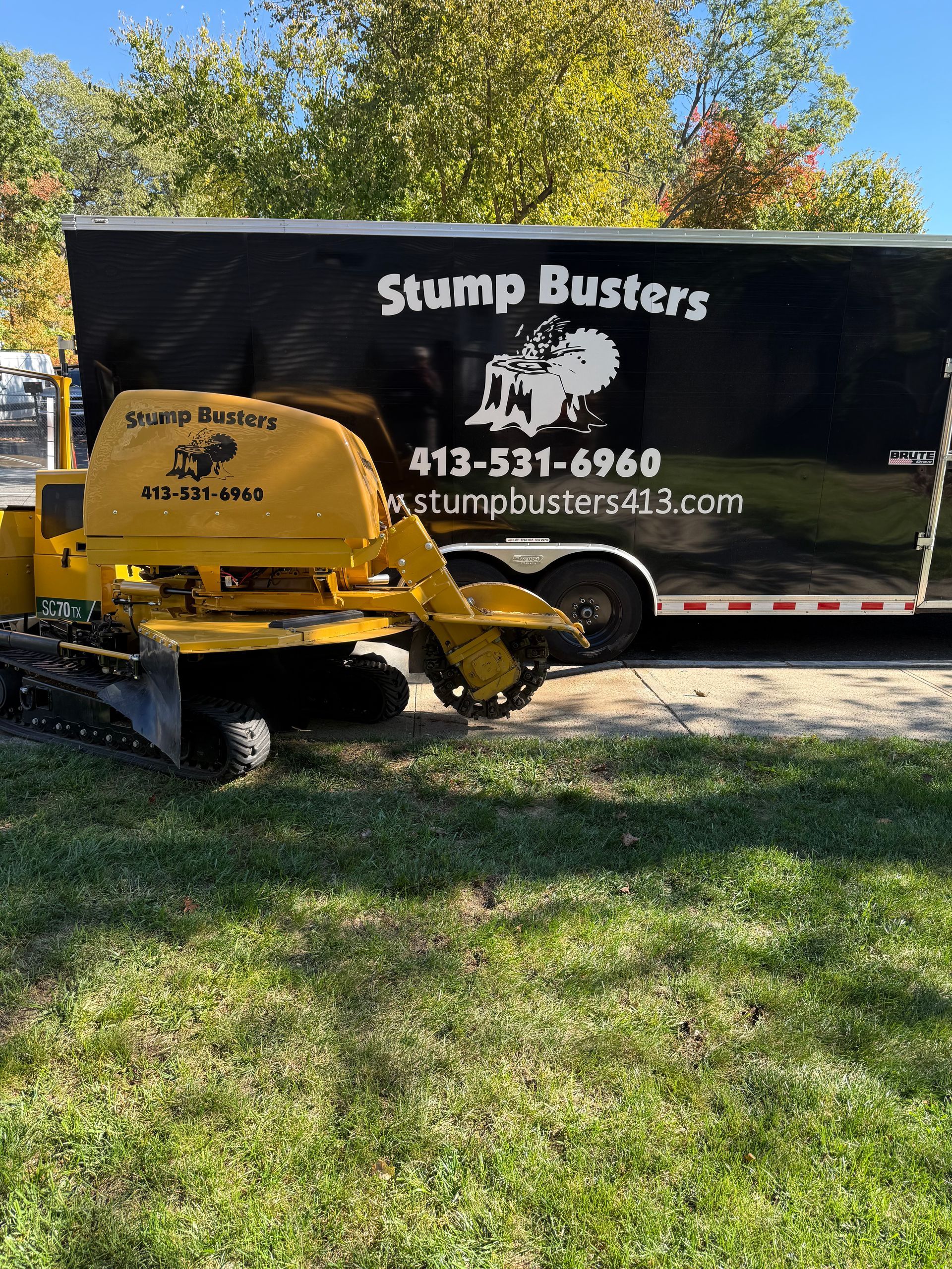 A Stump Busters stump grinder machine parked next to a black trailer with company logo and phone number.