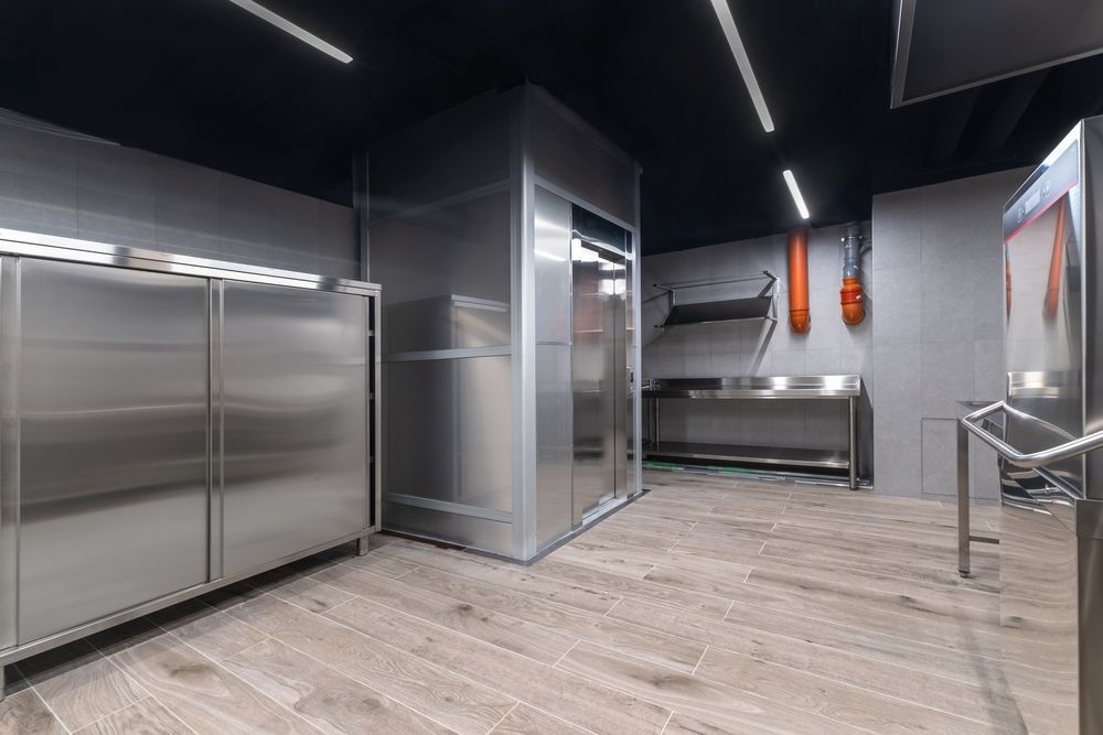 Modern stainless-steel commercial kitchen with bright overhead lights and light wood-look flooring