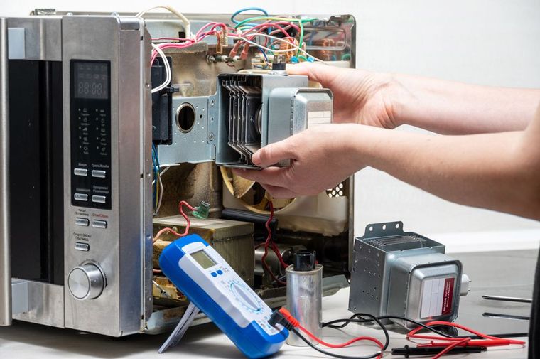 Hands working inside an open microwave with exposed wiring and components on a workbench