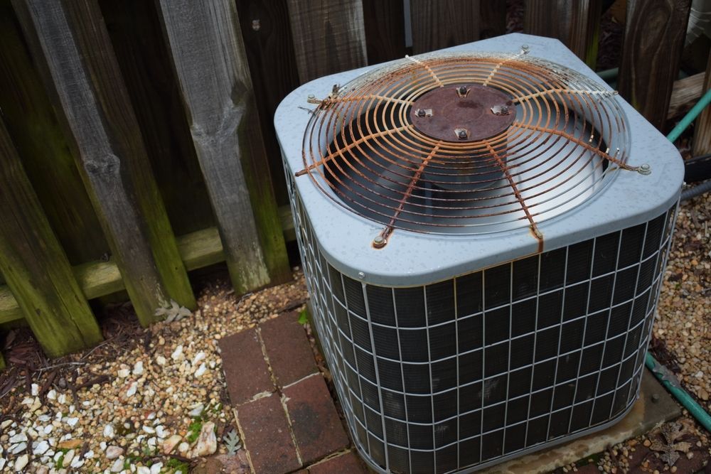 Outdoor air conditioner unit with a rusted fan top beside a wooden fence and brick pavers