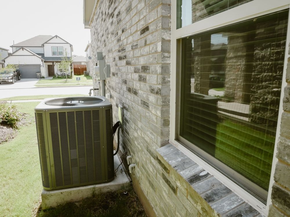 Outdoor air conditioner unit beside a brick house wall and window, with suburban homes in the background