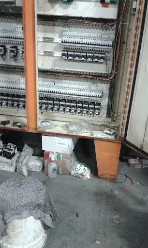 Open electrical control panel with rows of wiring and switches, scattered items on the floor below.