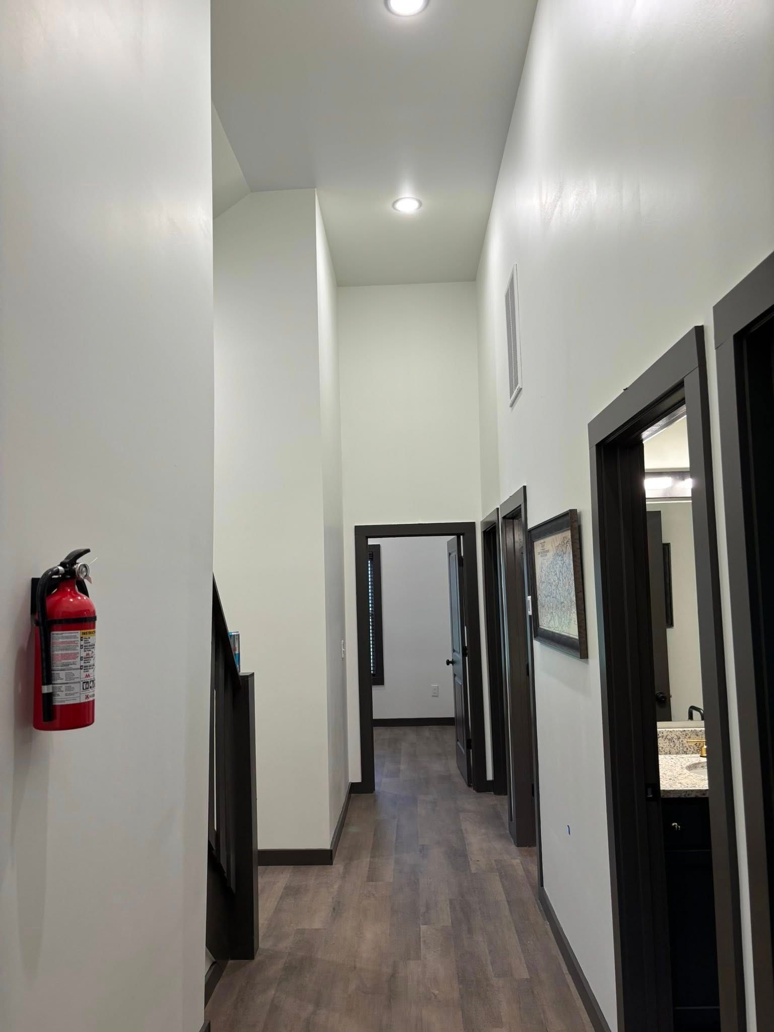 Narrow hallway with white walls, dark trim, wood floor, recessed lights, and a fire extinguisher on the left wall