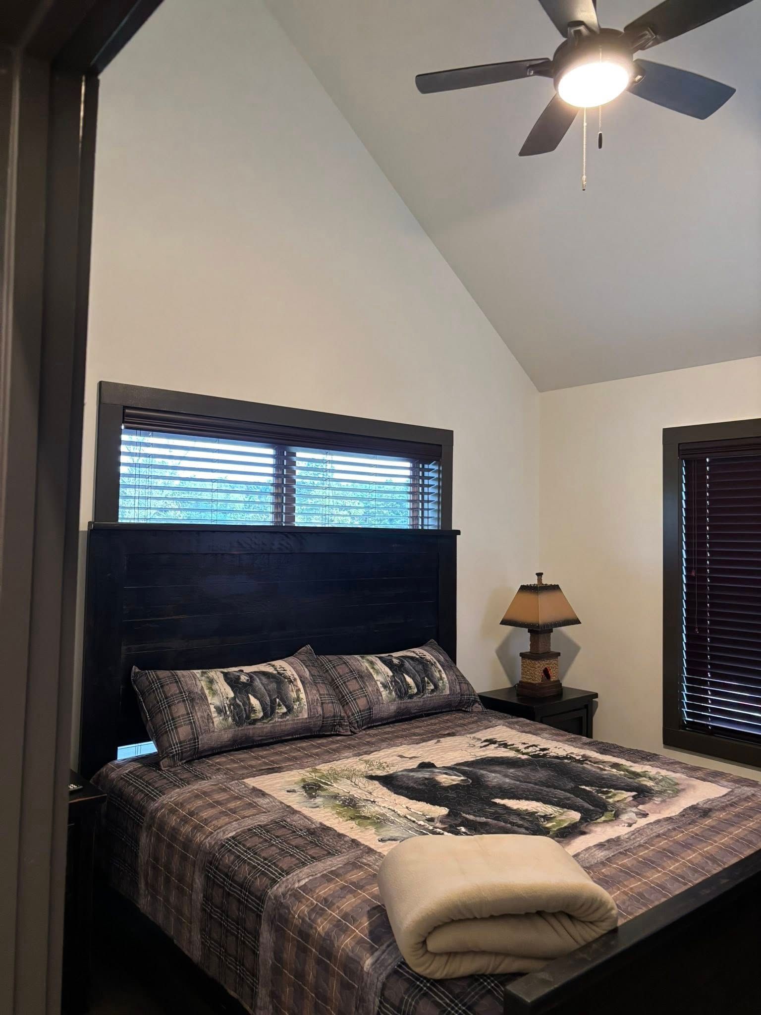 Bedroom with a dark wood bed, patterned comforter, folded towel, window blinds, lamp, and ceiling fan.