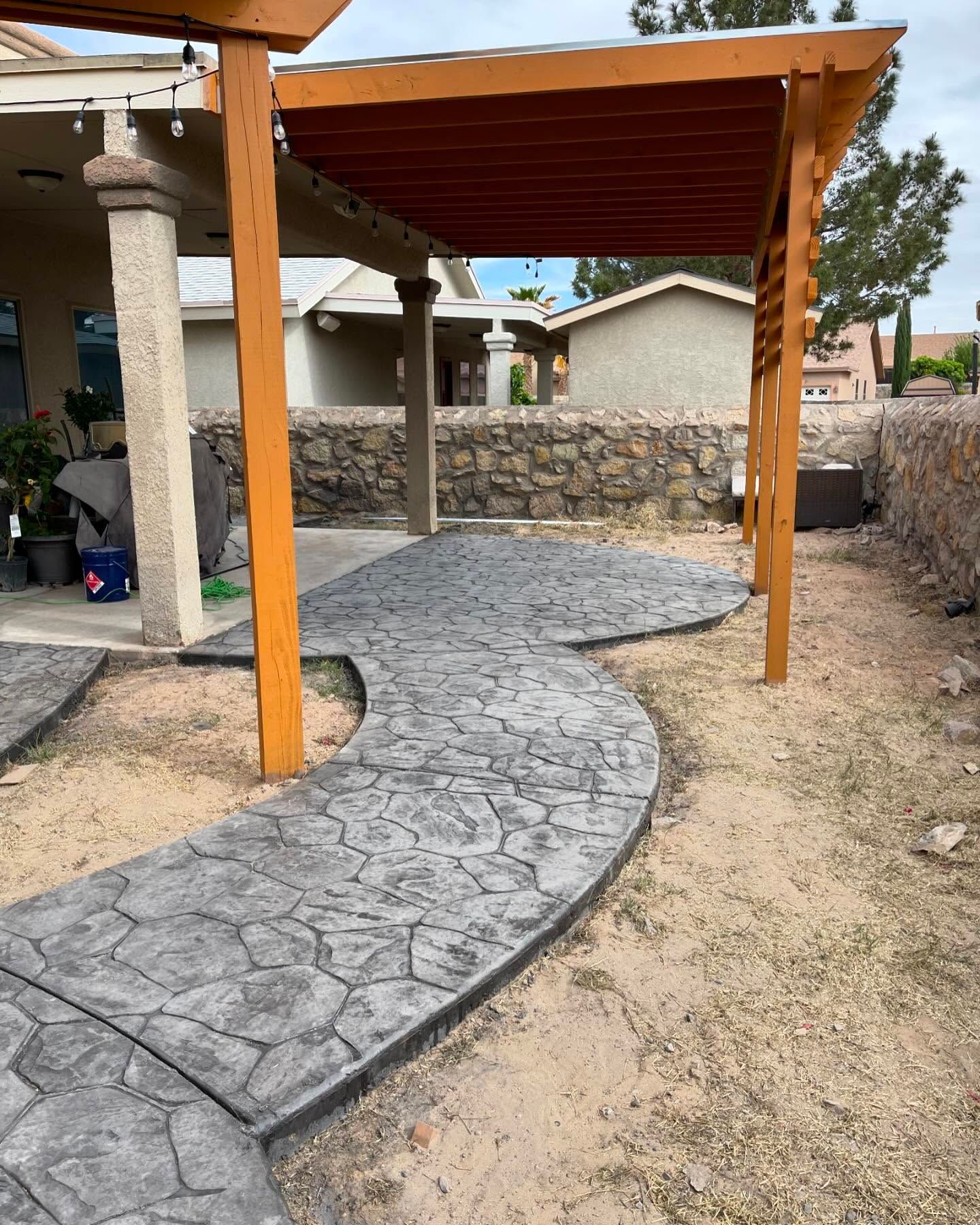 Curved stamped-concrete path under a wooden pergola in a dry backyard patio area.