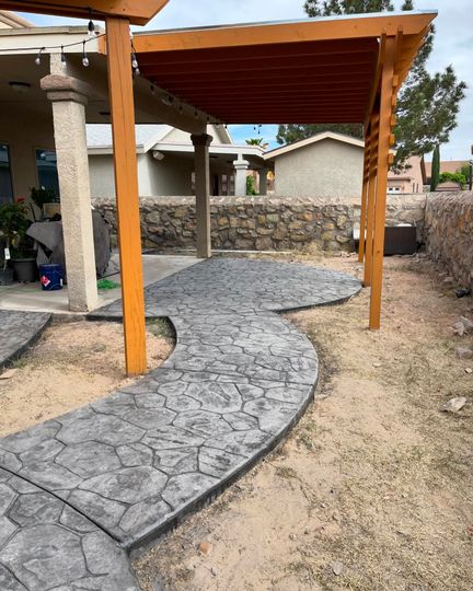 Curved stamped-concrete walkway under a wooden pergola in a desert-style yard, leading to a stone wall and house.