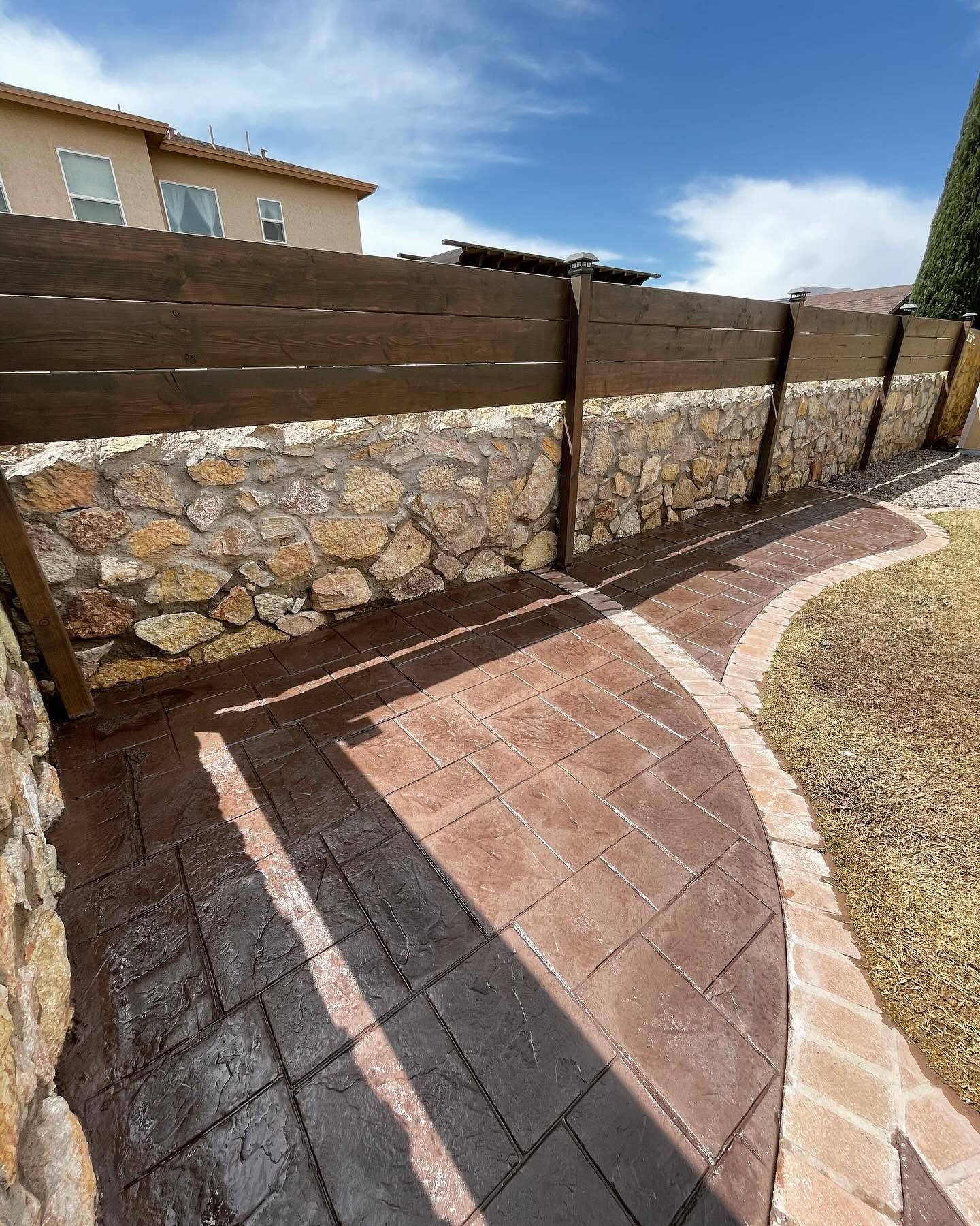 Stone and wood privacy fence beside a curved brick patio under a blue sky.