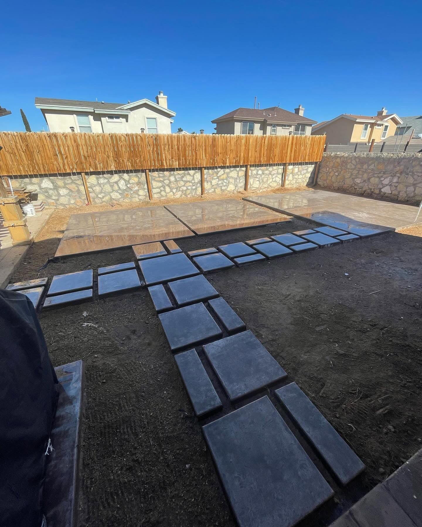 Backyard stone path under construction with dark pavers laid on dirt beside a wooden fence and houses.
