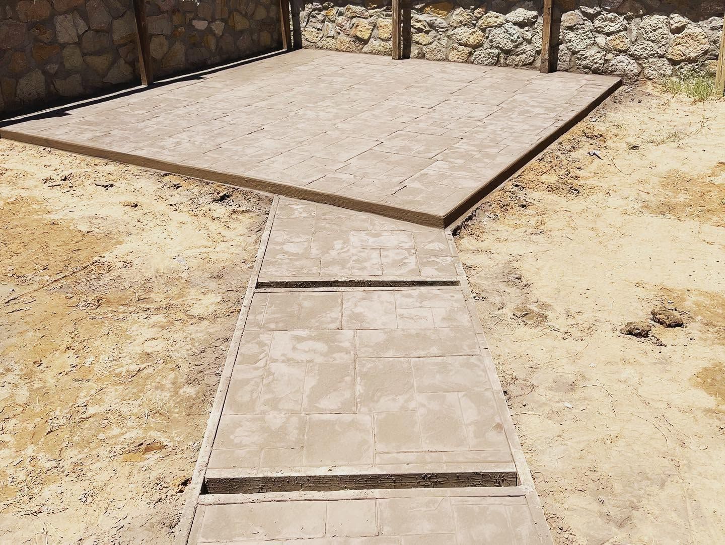 Concrete walkway leading to a raised patio slab in a sandy yard, with stone retaining walls.