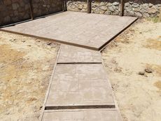 Concrete walkway leading to a raised patio slab in a sandy yard, with stone retaining walls.