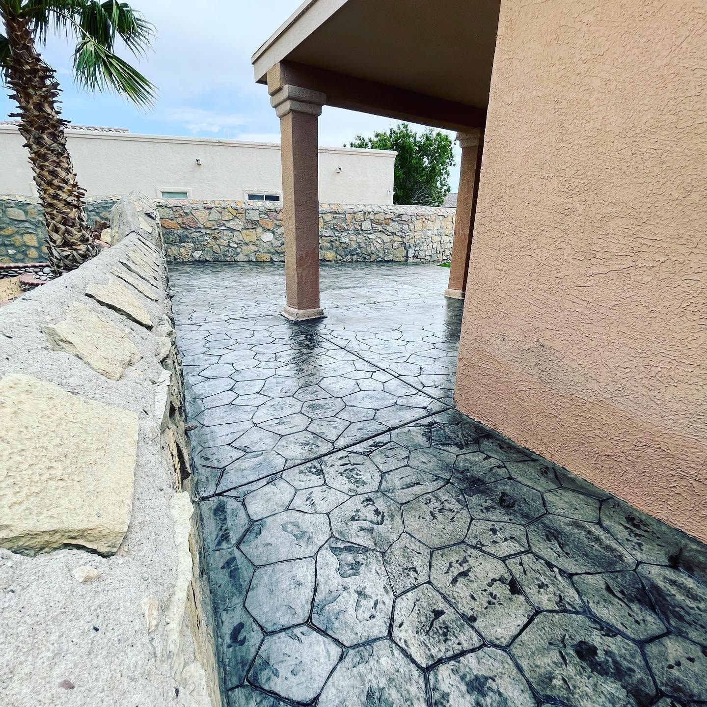 Stone patio and covered walkway beside a beige building, with a low stone wall and palm tree.
