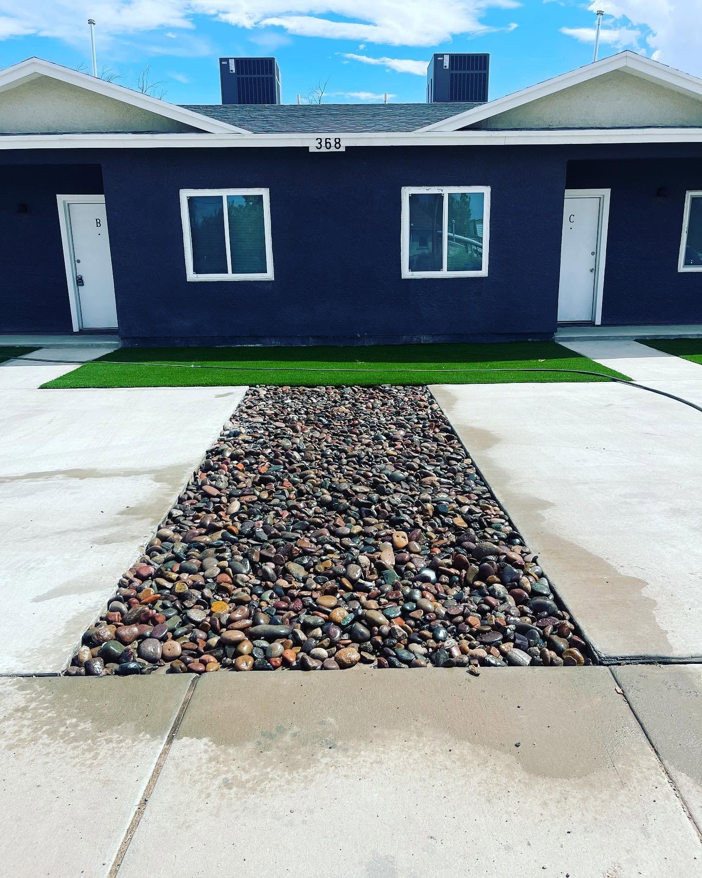 Modern duplex with blue exterior, white doors, and a rock-lined walkway between concrete driveways.