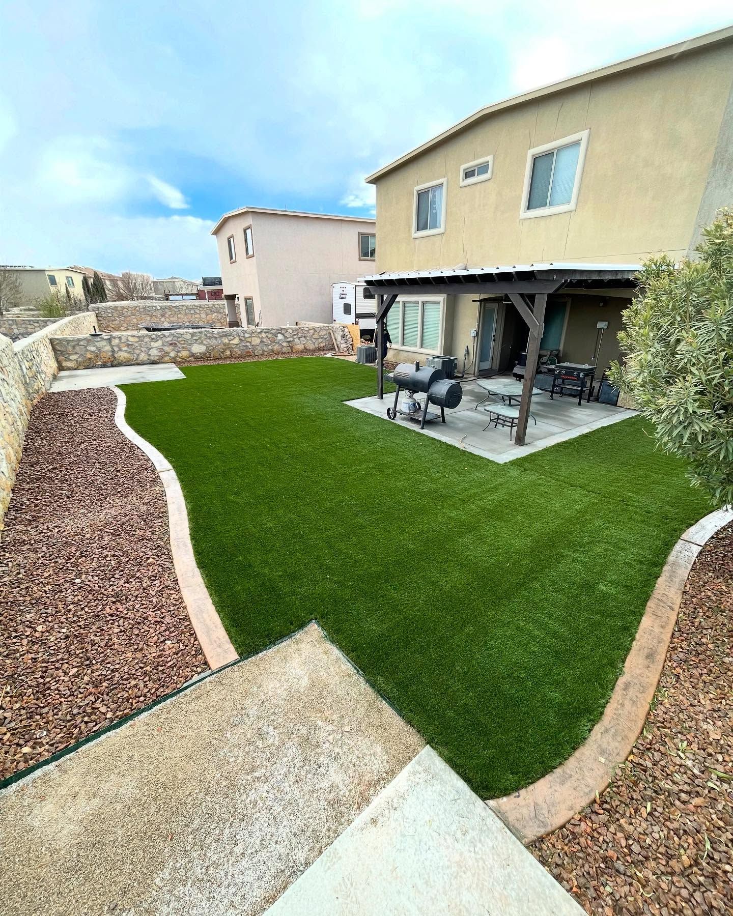Modern backyard patio with artificial grass, stone borders, and outdoor seating beside a beige house