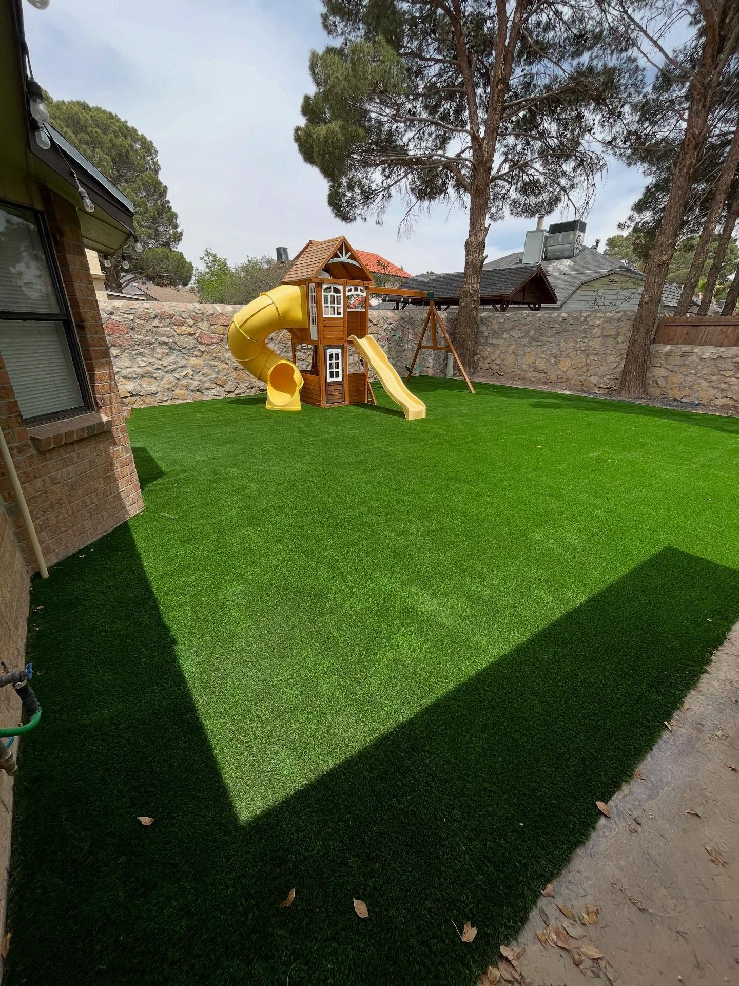 Backyard playground with green turf, a yellow slide, and stone walls under a bright sky