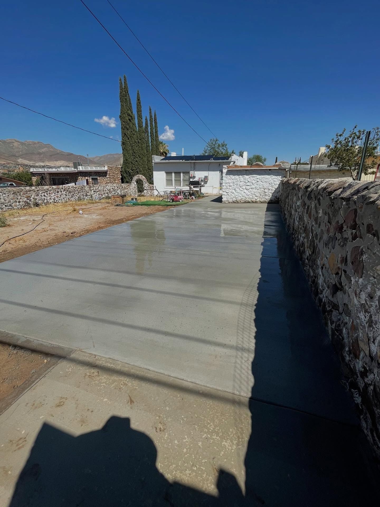 Concrete driveway beside a stone wall and house under a clear blue sky, with trees and mountains in the distance