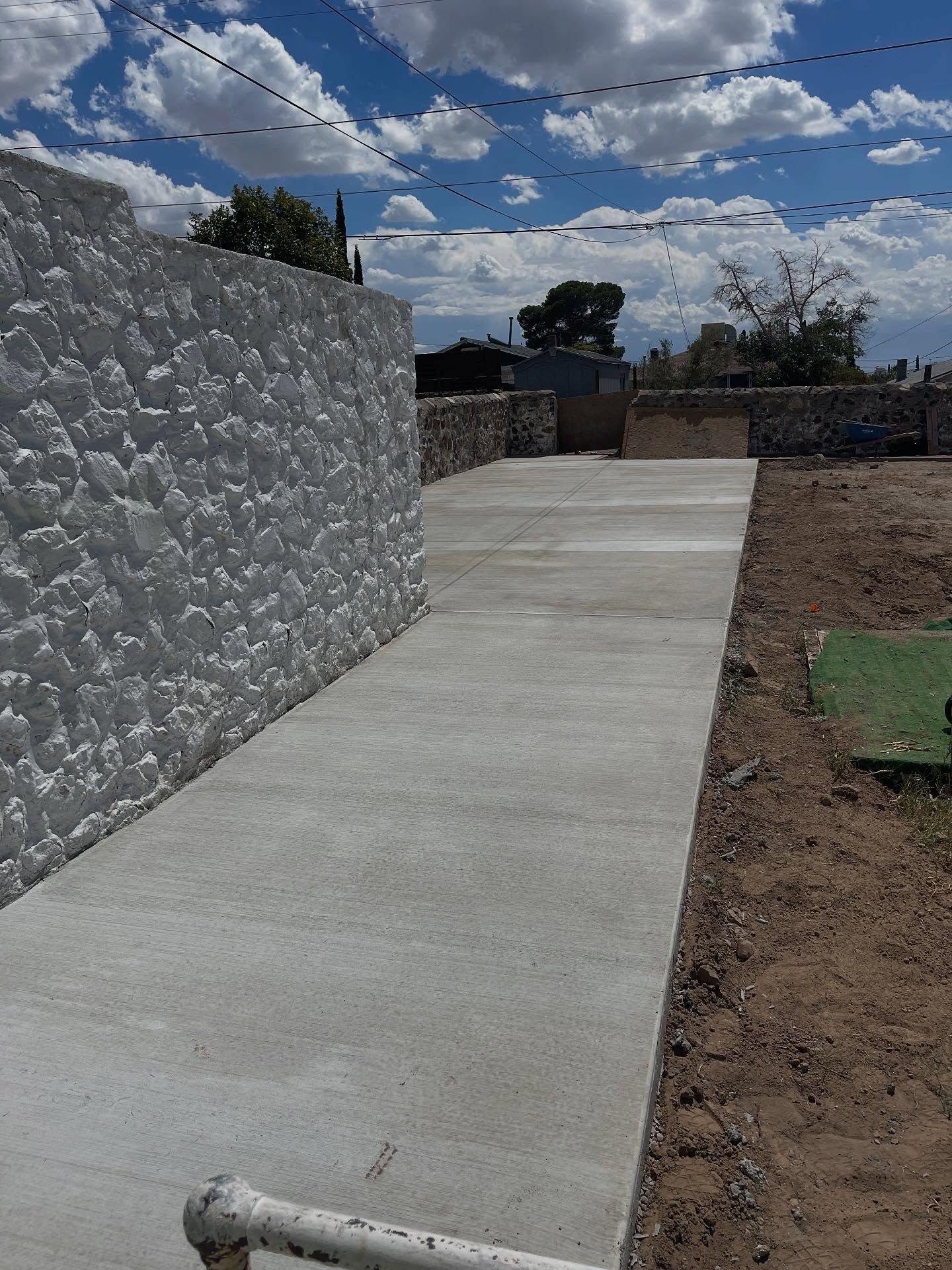 Concrete walkway beside a white textured wall under a cloudy sky in a backyard.