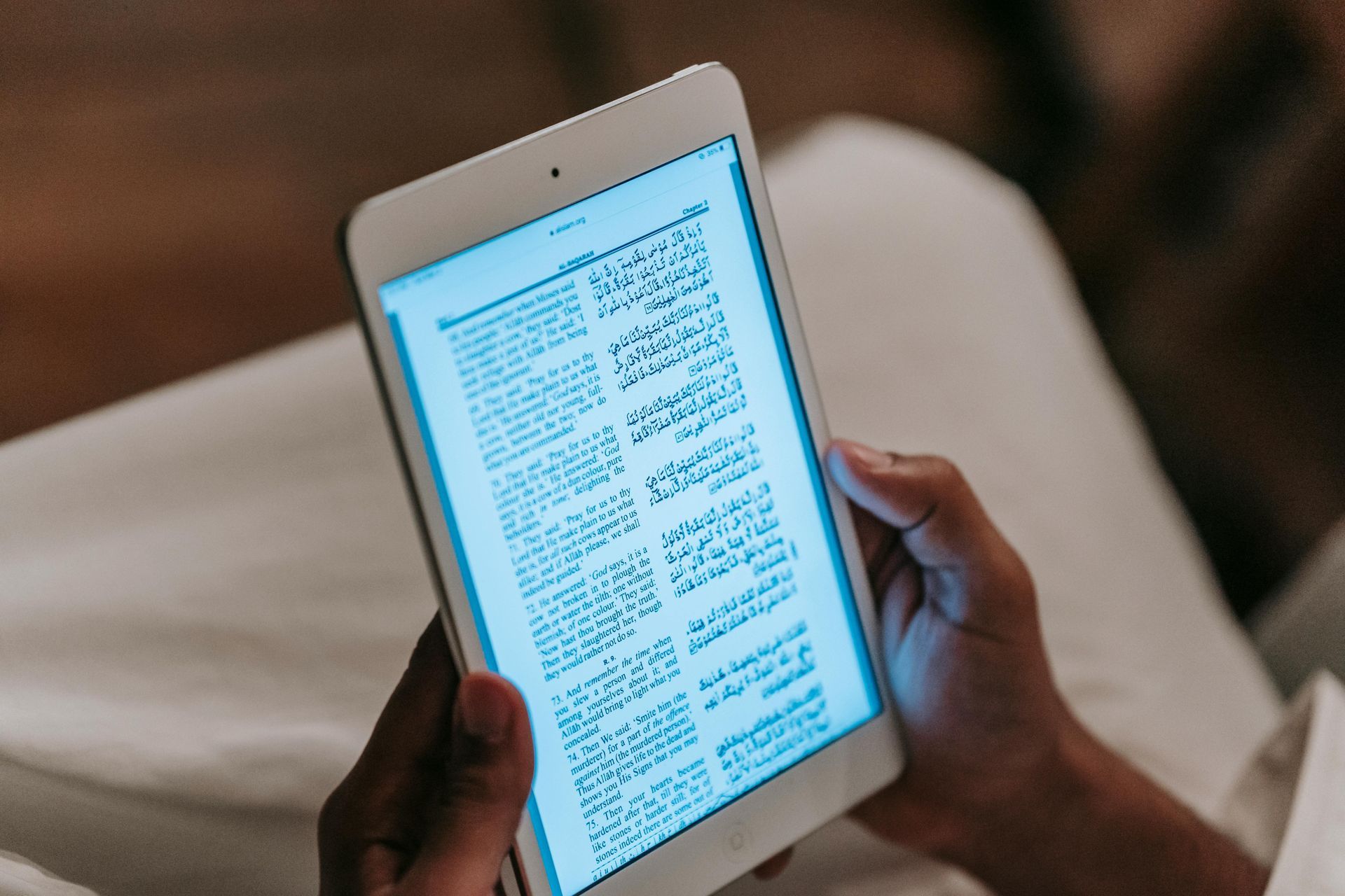 Hands holding a white e-reader displaying a text page in a dim indoor setting