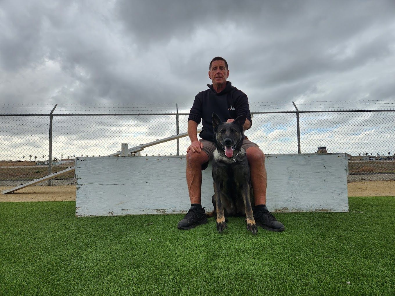 Rich Rosen, owner of Dockside Dog Training, on a concrete barrier with dog on green turf; chain-link fence, cloudy sky.