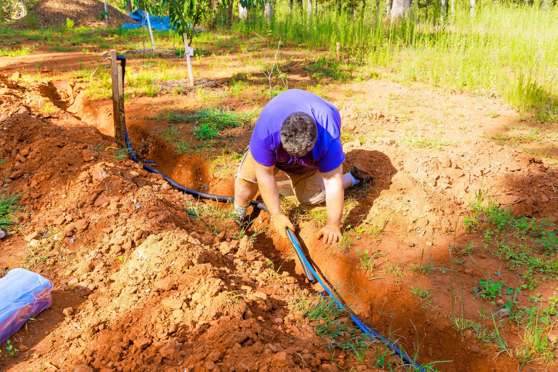 Person in a blue shirt crouches in a garden, digging soil beside a black irrigation hose.