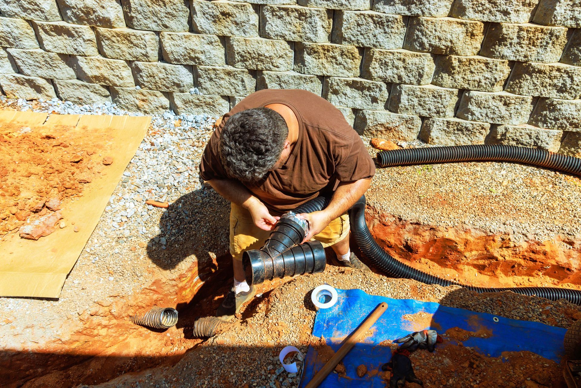 Worker connecting a large black pipe beside a stone wall, with tools and blue tarp nearby.