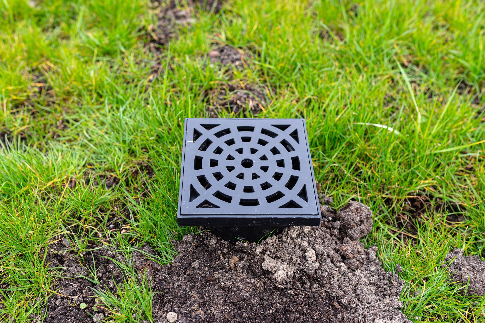 Black drainage grate set in a grassy patch with exposed soil around it