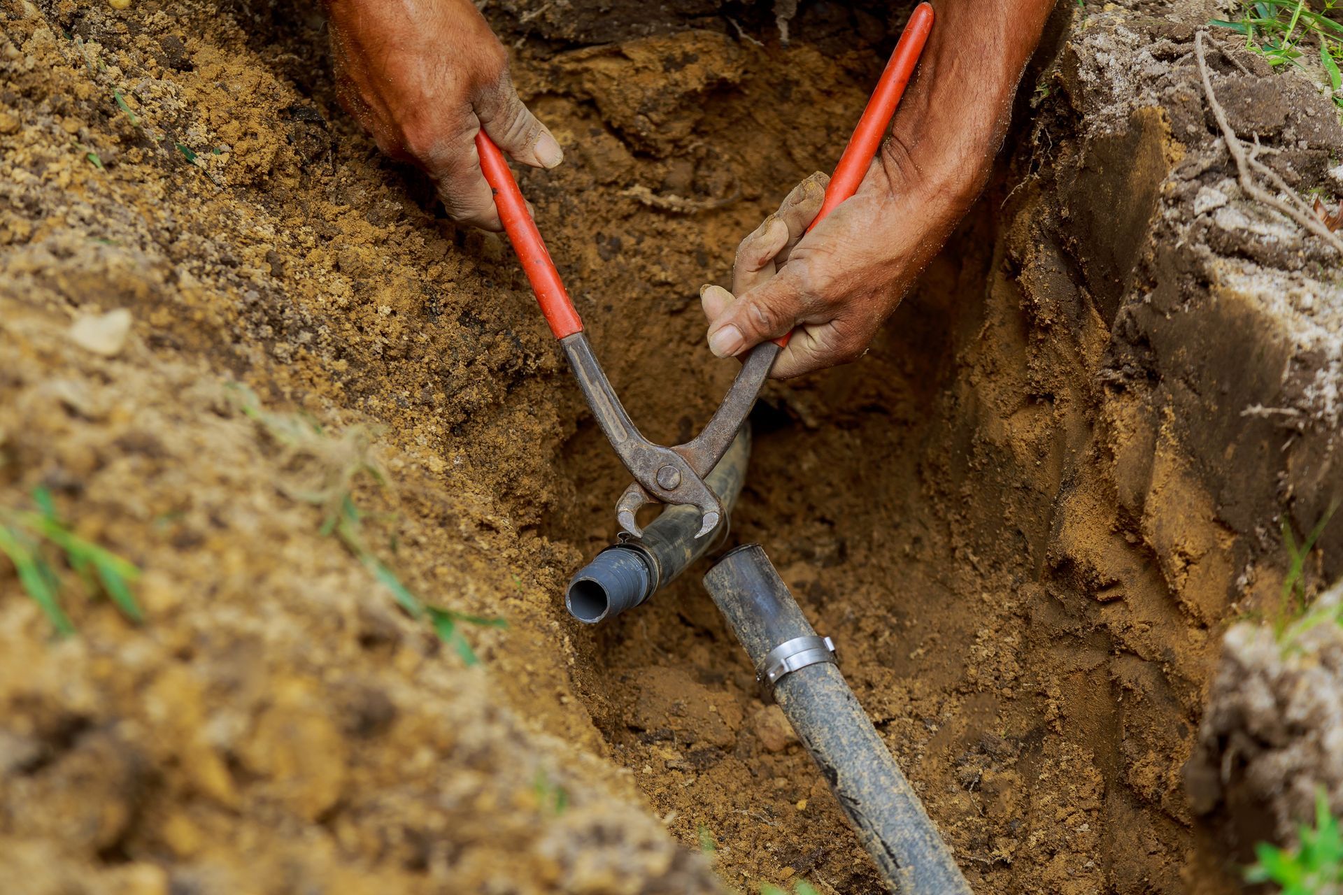 Hands using pliers to repair a black pipe in a dirt trench
