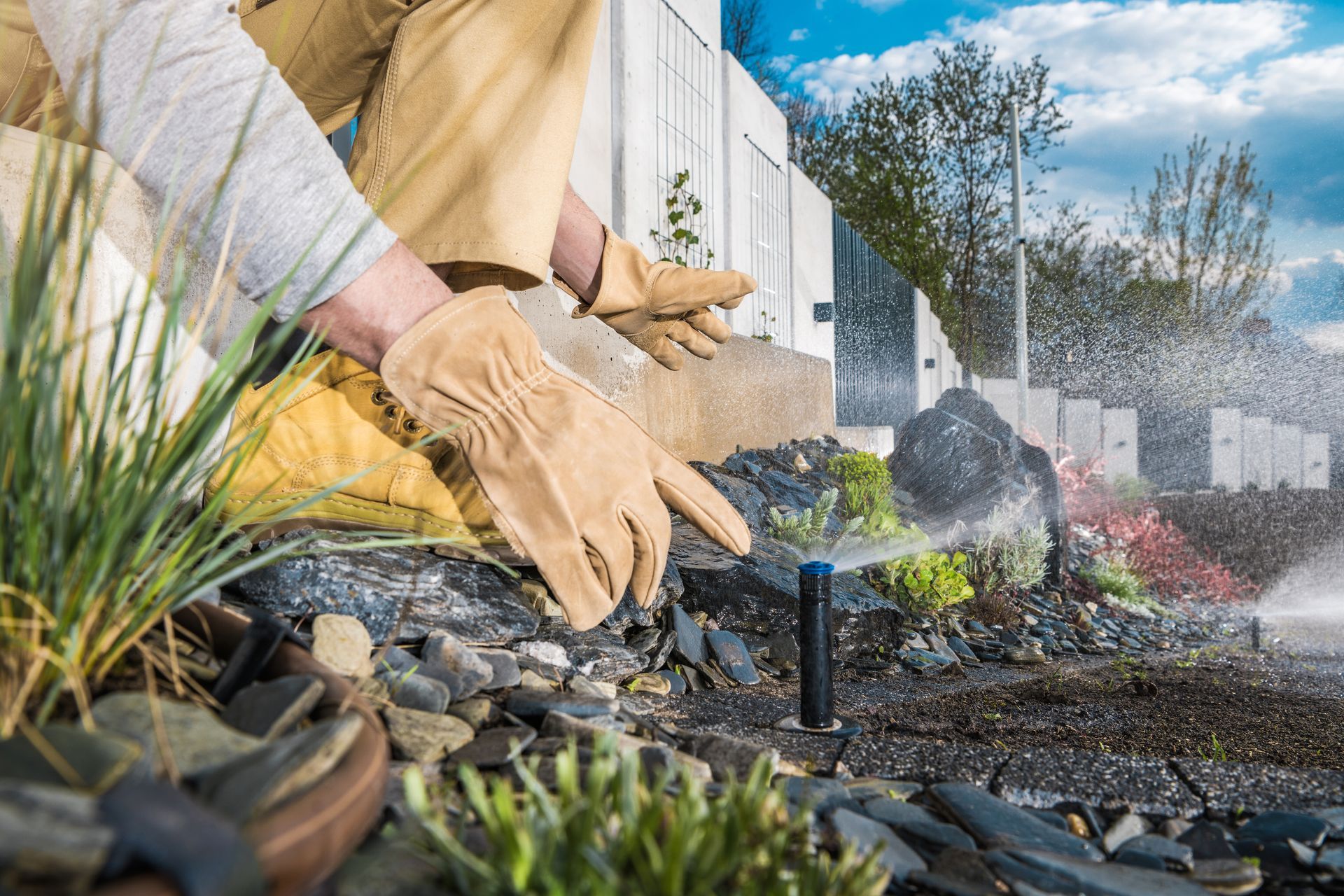 Gloved hands spraying water over a garden bed with rocks and plants beside a building