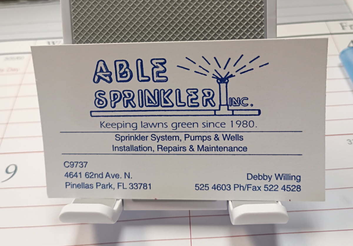 Able Sprinkler Inc. business card on a holder, white card with blue logo and text.