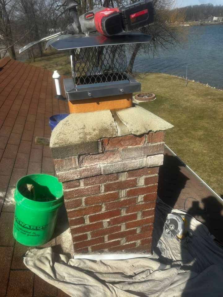 Brick chimney with a black-and-red power tool on top, beside a green bucket and a lakeside deck.