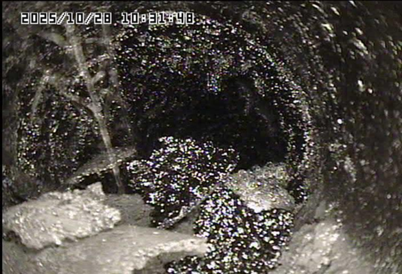 Inside a dark pipe, muddy water and debris flow around a rough, wet tunnel interior.