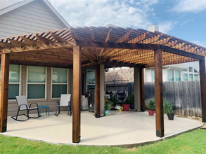 Wooden pergola over a concrete patio with a house in the background and green grass. Two chairs sit on the patio.