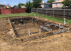 Foundation layout marked with rebar and white stakes on a dirt lot in a fenced backyard.