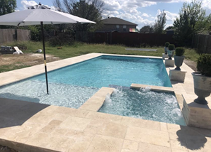 Backyard swimming pool with a shallow ledge, umbrella, and stone patio under a blue sky