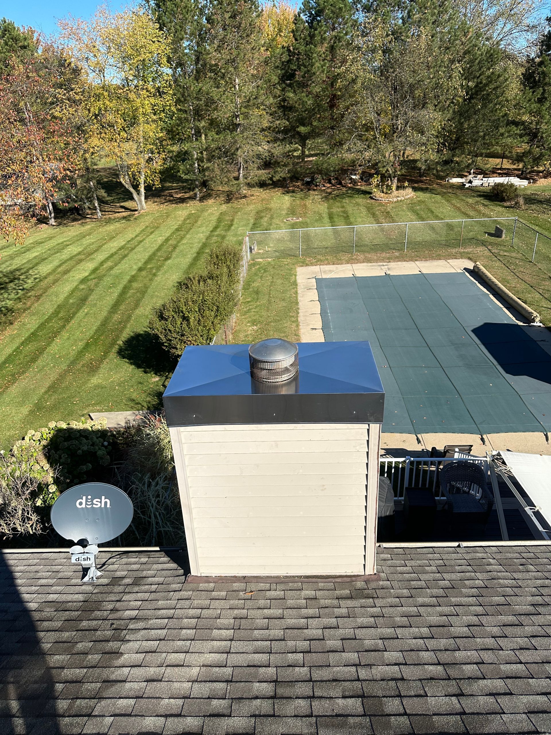 Backyard view from a roof with chimney, satellite dish, grassy lawn, and trees beyond