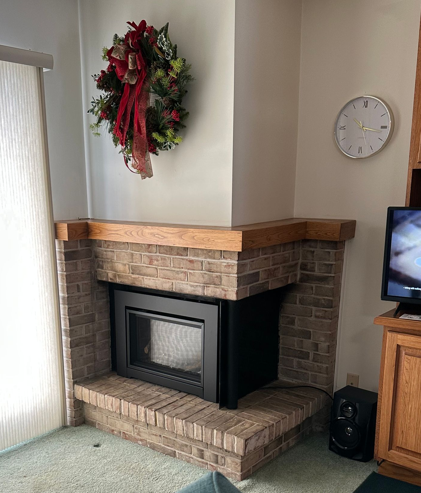 Living room corner with brick fireplace, wreath, wall clock, and TV on a wooden stand