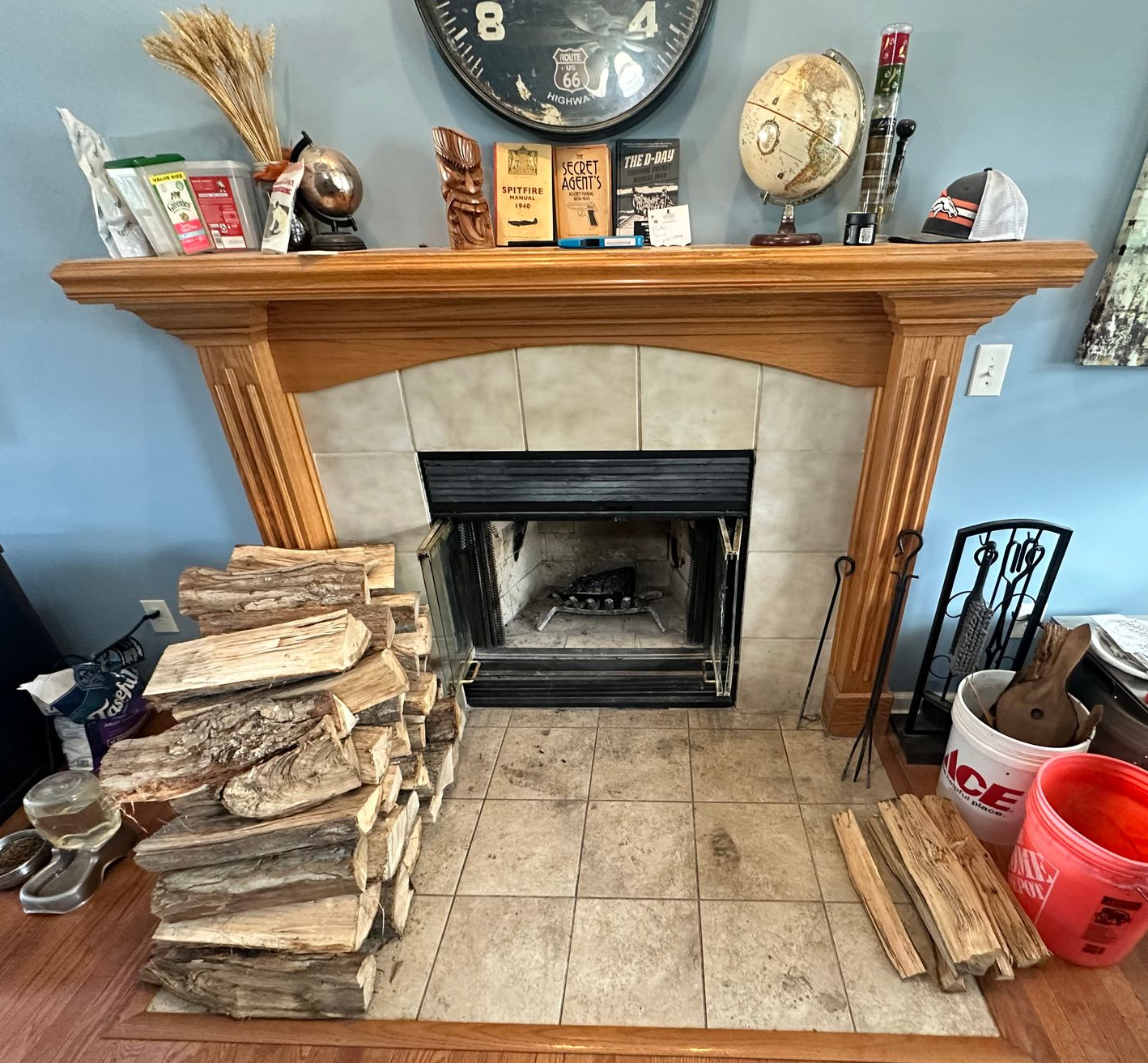 Brick fireplace with wooden mantel, stacked firewood, and decorative items in a tiled living room.