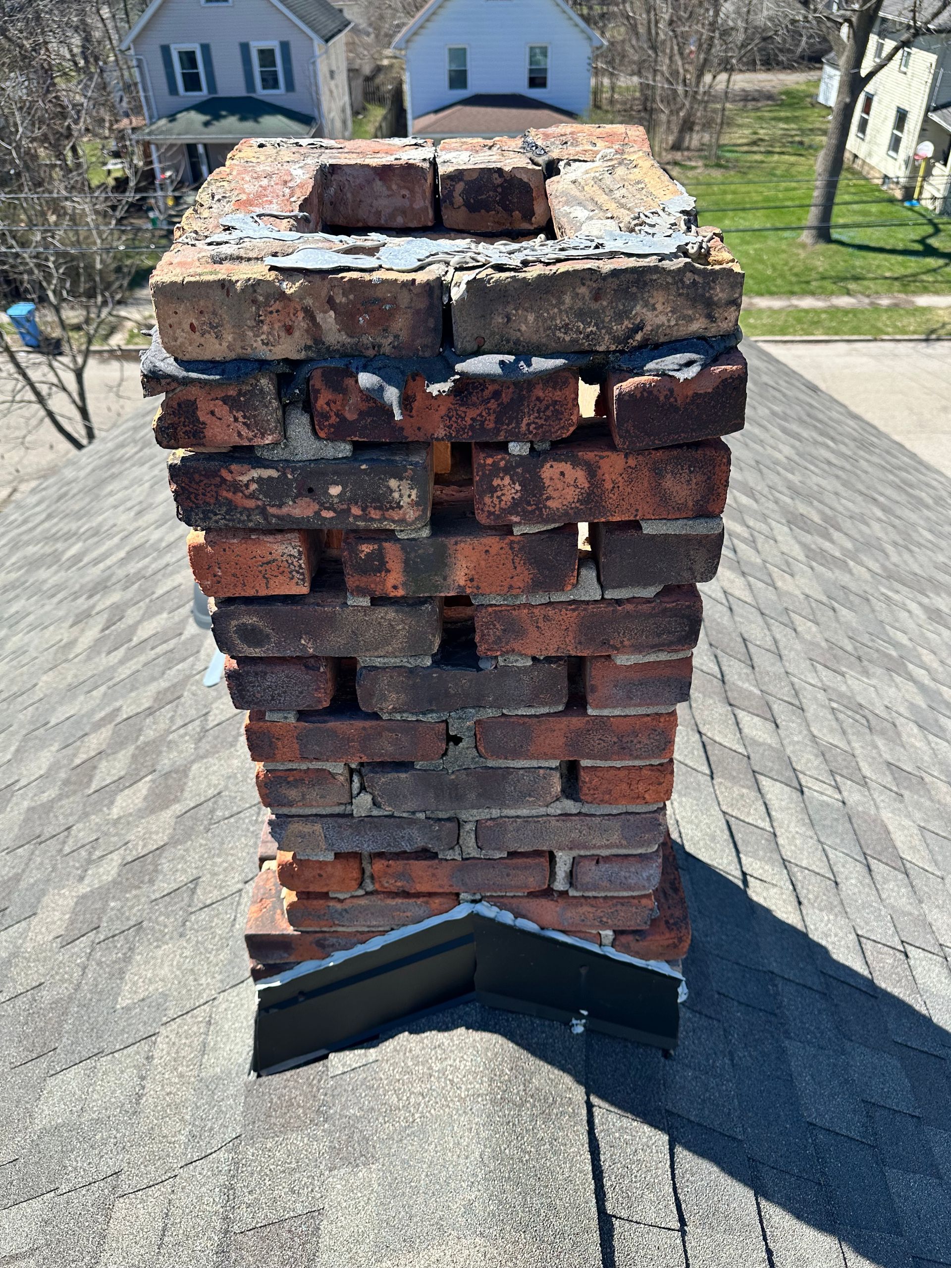 Damaged brick chimney with missing top bricks on a shingled roof, viewed from above.