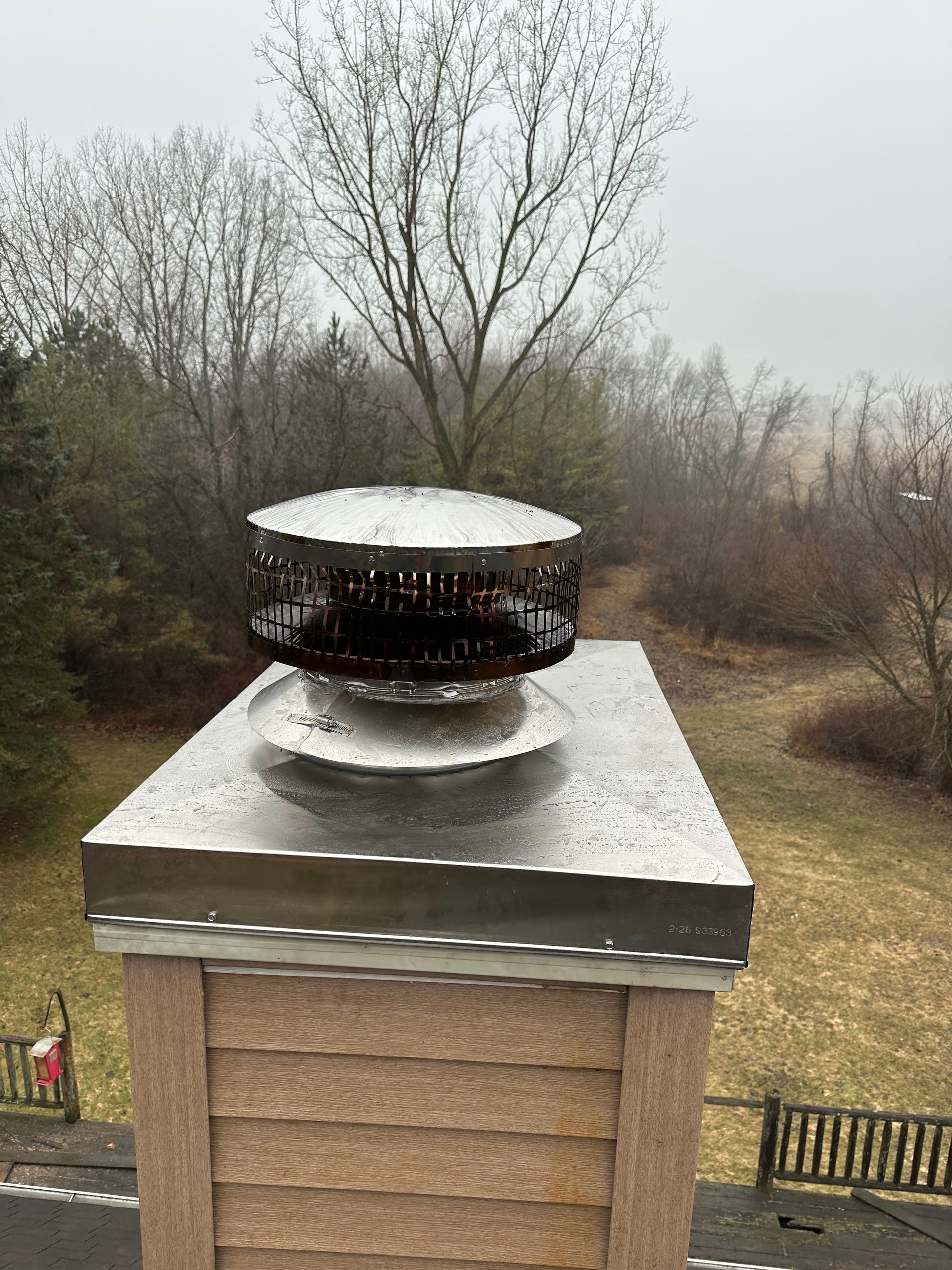 Rooftop vent on a brick chimney with snow on top, surrounded by leafless trees.