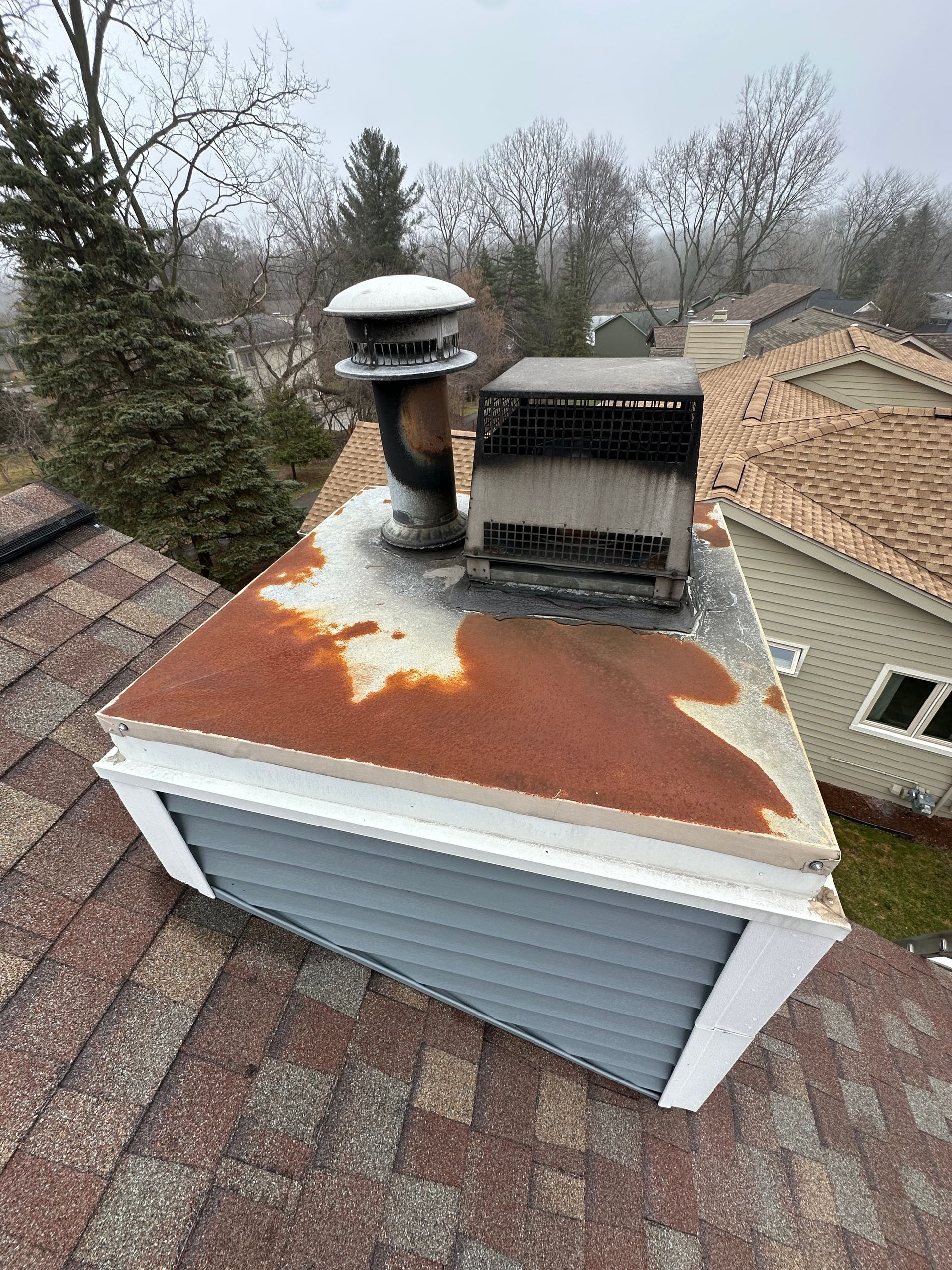 Rusty rooftop vent and HVAC unit on a small roof, with trees and neighboring houses in the background