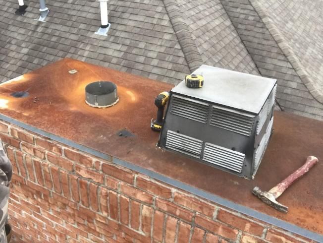 Rusted flat roof with brick chimney. A vent cover, tools, and metal cap are visible. A roof is in the background.