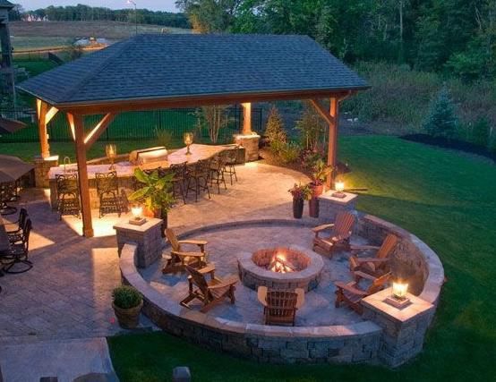 Outdoor patio with fire pit, bar, seating under a wooden roof, all illuminated at dusk.