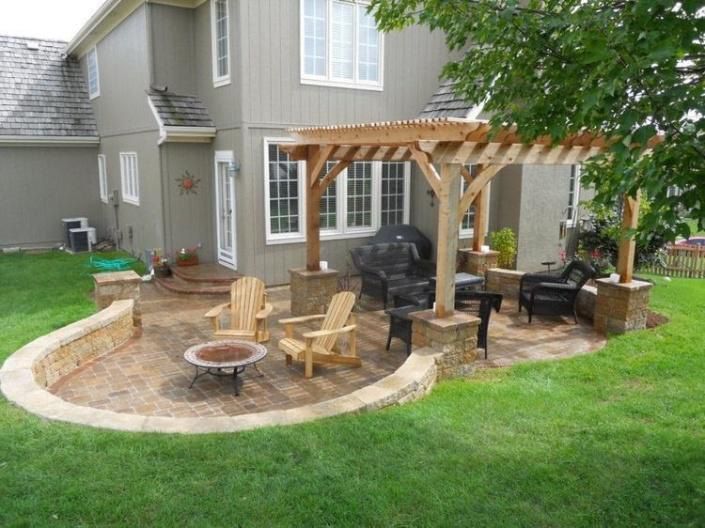 Backyard patio with wooden pergola, seating, fire pit, and low retaining wall; next to a house with green grass.