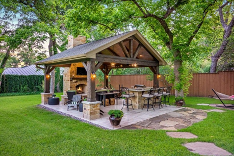 Covered backyard patio with dining table, seating, and stone fire pit on a green lawn under trees
