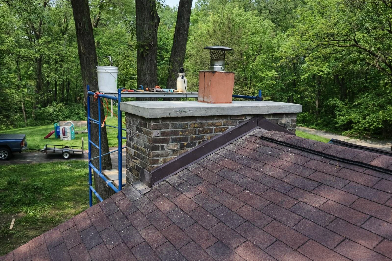 Roof and chimney on a house amid green trees, with a ladder beside the chimney.