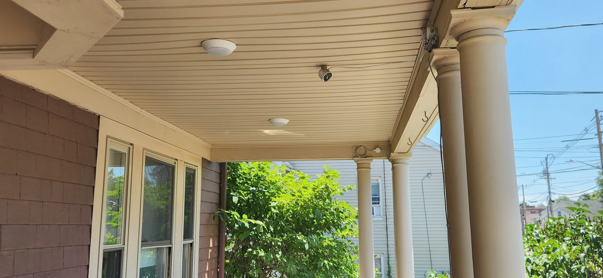 Covered front porch with white columns, beige siding, and windows beside green shrubs under a clear blue sky