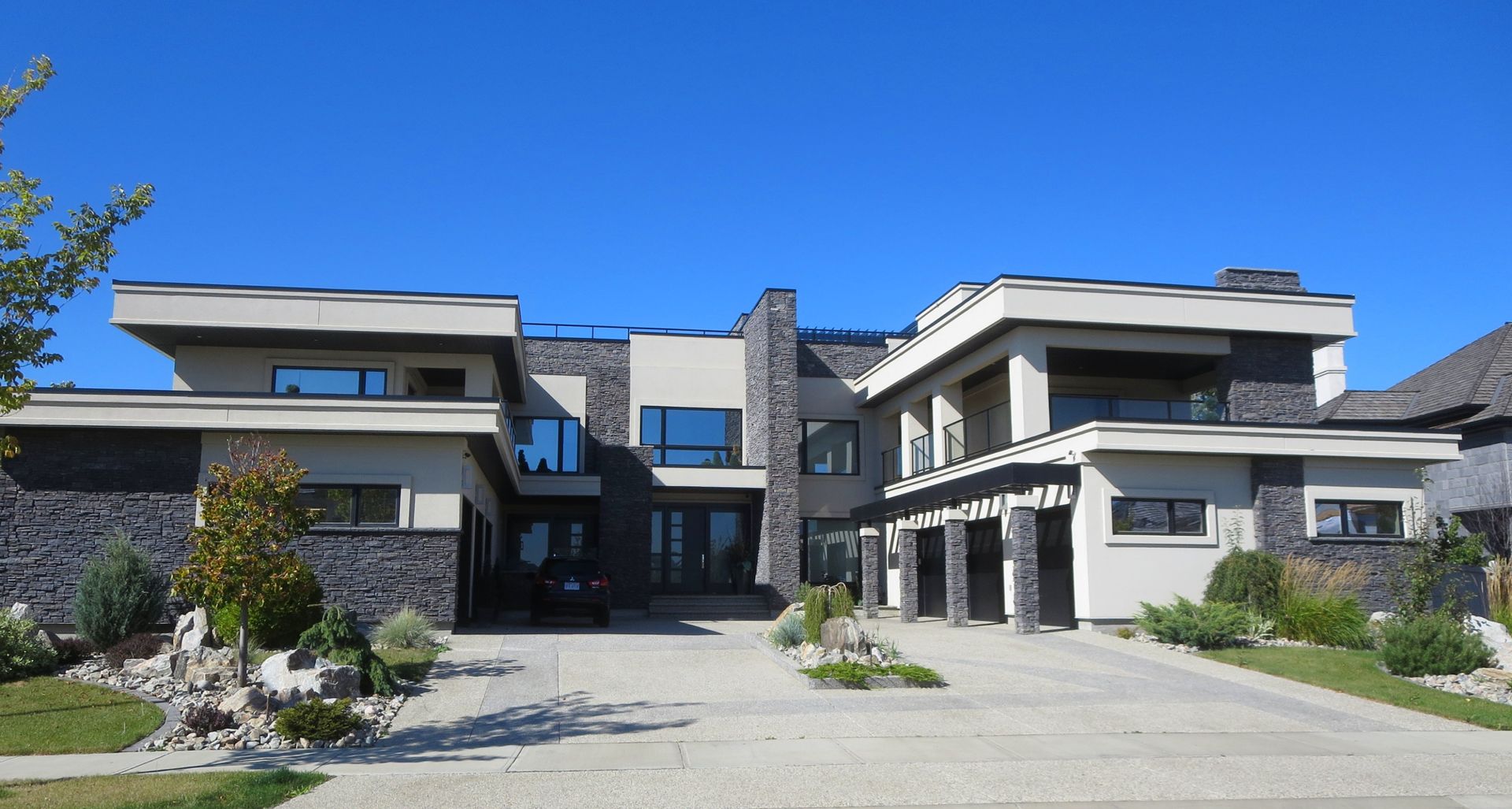 Modern two-story house with stone and tan exterior, blue sky, gravel driveway.
