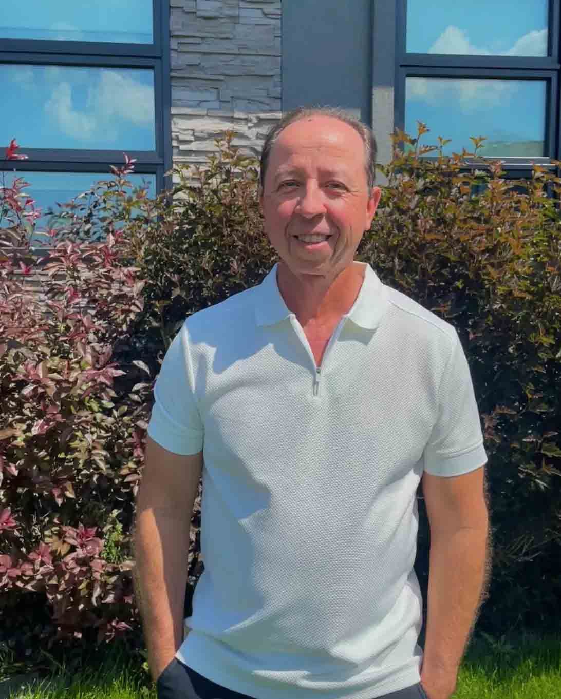 Man in white polo shirt smiles, standing outdoors in front of a building with windows, and shrubbery.
