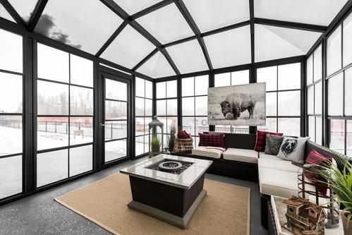 Sunroom with black-framed windows, beige rug, sectional sofa, fire pit, and a bison painting. Snow outside.