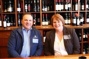 A man and a woman are sitting at a table in front of shelves of wine bottles.
