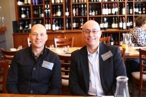 Two men are sitting at a table in front of a wine shelf.